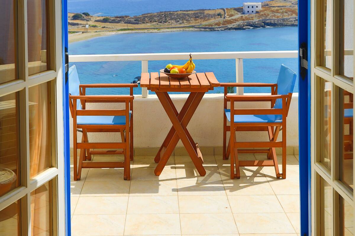 Een open balkon met een tafel en stoelen, een schaal fruit en uitzicht op een kustlandschap met een wit gebouw.