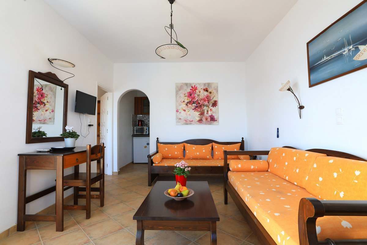 A living room with a sofa set, a dining table with fruit, a TV, and artwork on the walls.