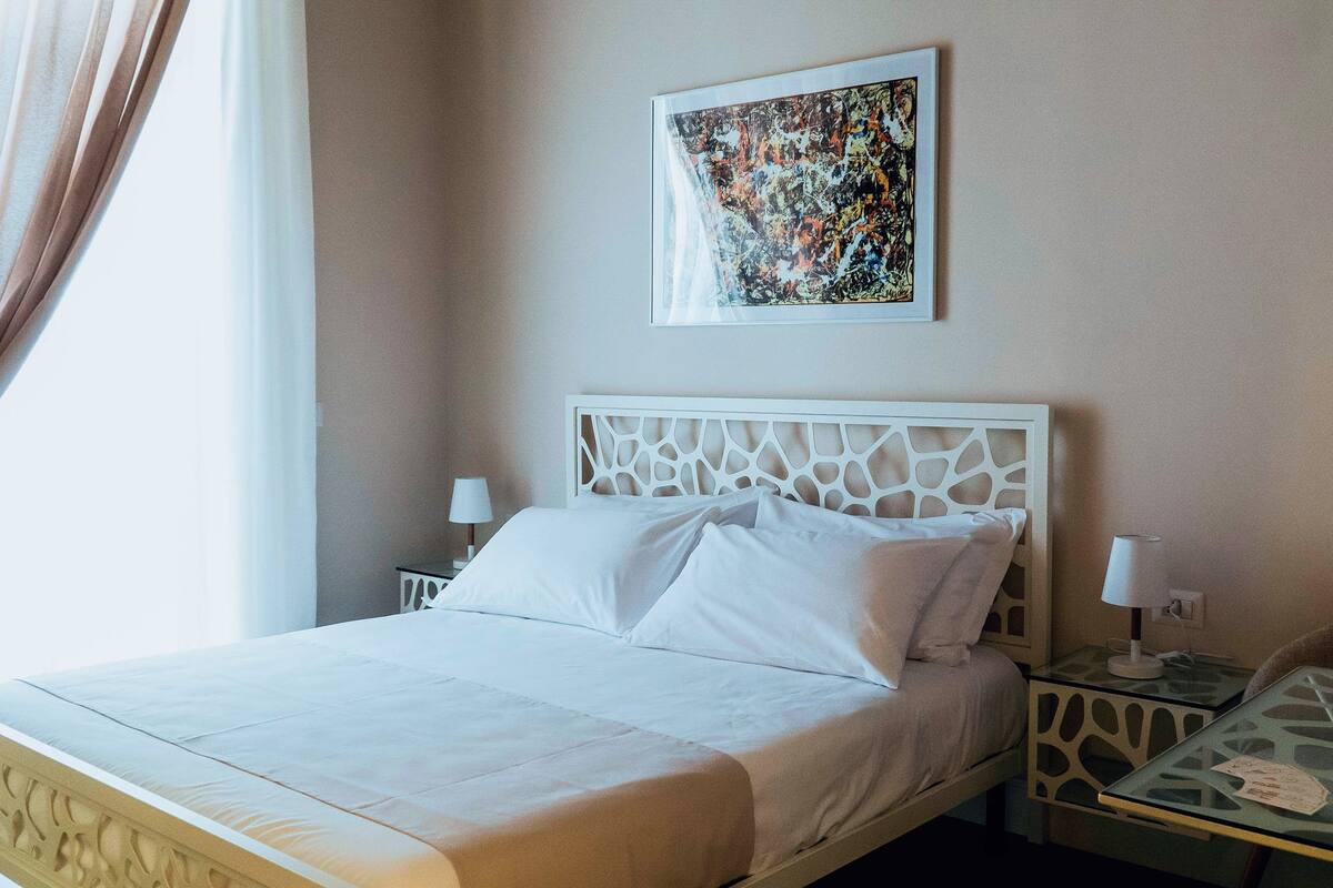 Ein Hotelzimmer mit einem Bett, zwei Nachttischen mit Lampen, einem an der Wand hängenden abstrakten Gemälde und einem Fenster mit Vorhängen.