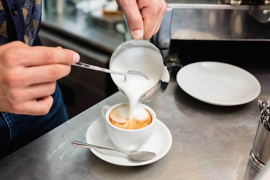 Eine Person gießt Milch in eine Kaffeetasse auf einem Edelstahl-Tisch.
