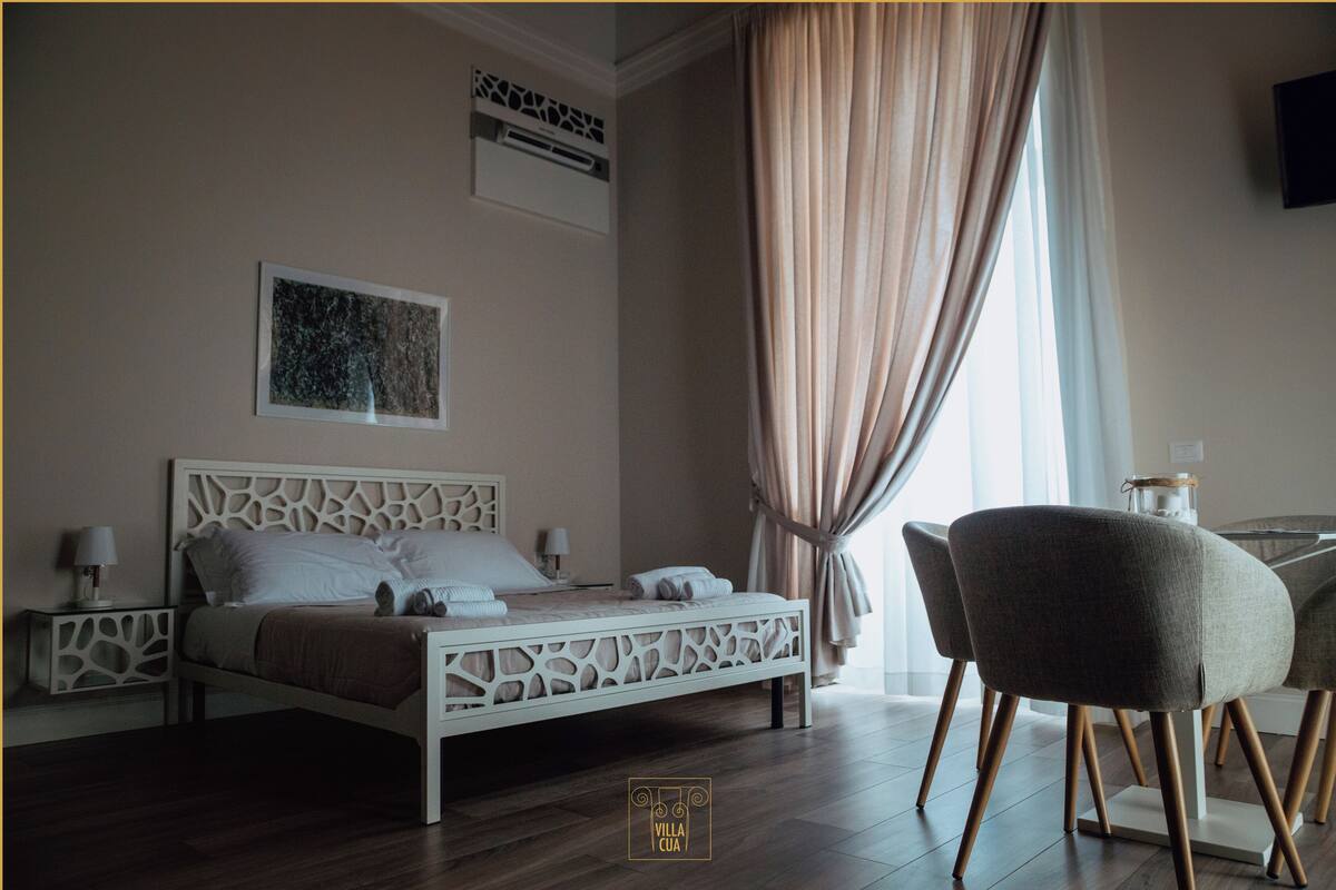 Ein Schlafzimmer mit einem weißen Bett, einem Nachttisch, einem Sessel und einem Fenster mit Vorhängen.