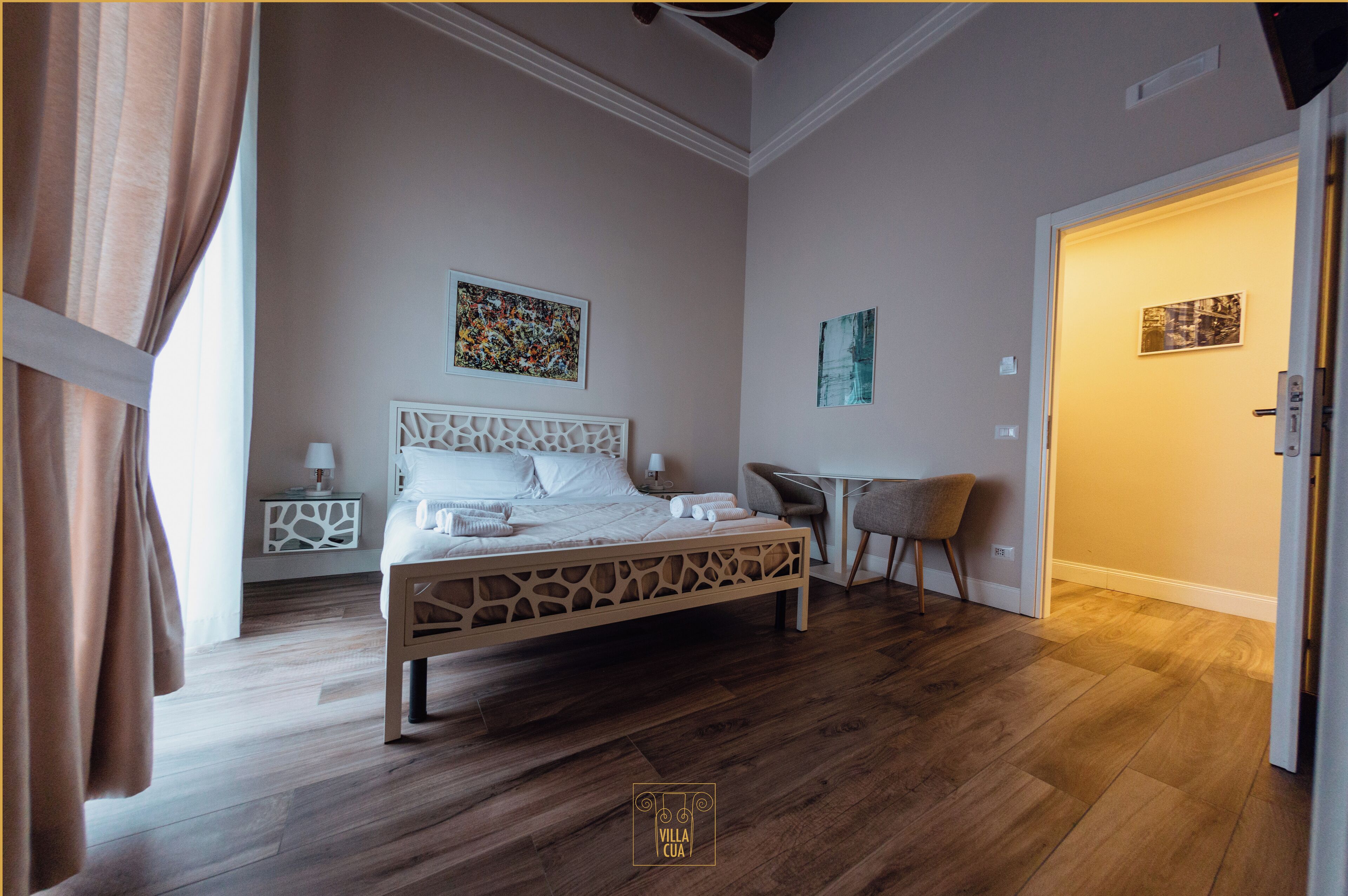 A bedroom with a wooden bed, a desk, and two chairs. There are two paintings on the wall and a door leading to another room.