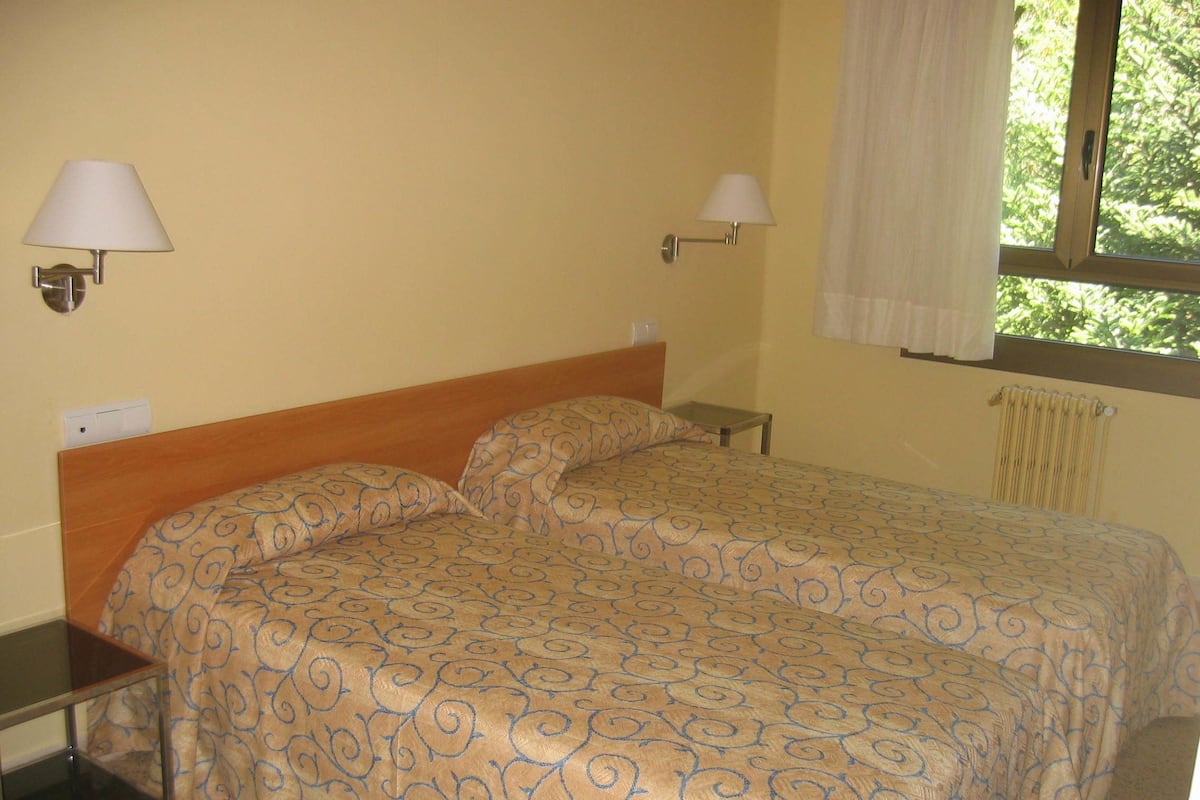 Una cama individual con colcha estampada, dos mesitas de noche, dos lámparas de pared, una ventana con cortina y un radiador.