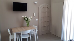 A dining area with a wooden table and white chairs, a vase with white flowers, a flat-screen TV mounted on the wall, and a door with a classic design. - HOLIDAY HOUSE MANFREDI HOME (Polignano a Mare)