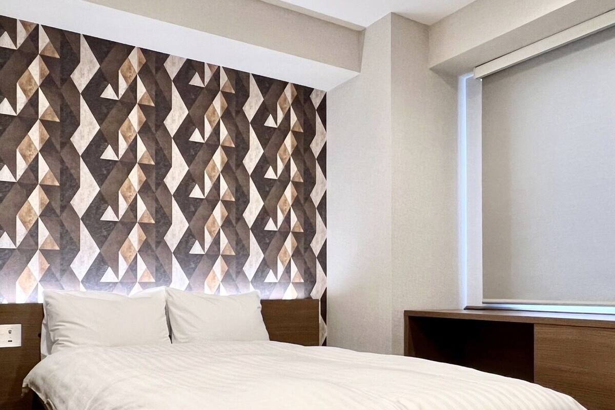 Una cama bien hecha con un cabecero de estampado geométrico, una mesita de noche de madera y una ventana con persianas.