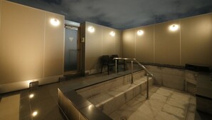 Un baño público moderno y bien iluminado, con separador de vidrio, baranda metálica y un banco.