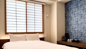 Habitación de hotel con una cama grande, una ventana con persianas, una mesita de noche con cafetera y una pared azul con textura.