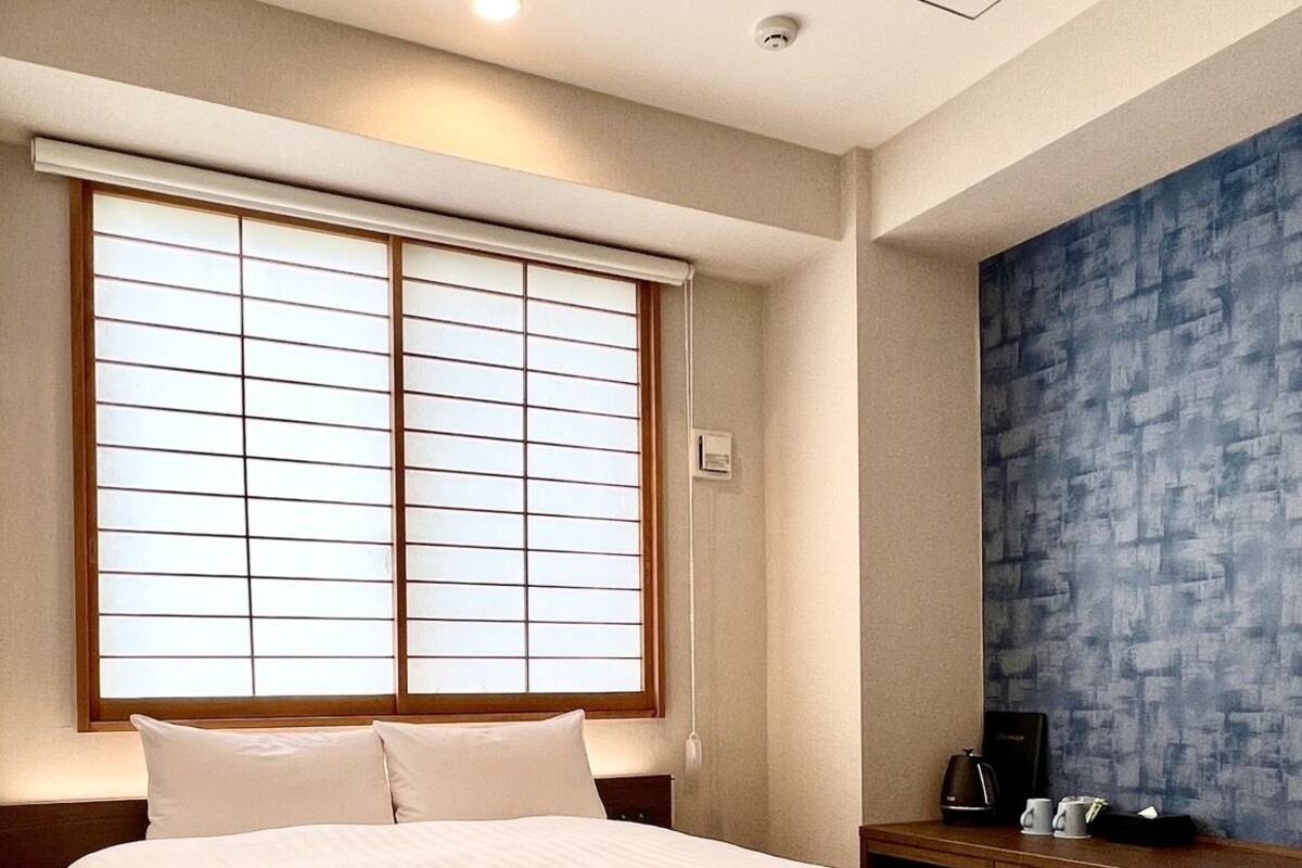 Habitación de hotel con una cama grande, una ventana con persianas, una mesita de noche con cafetera y una pared azul con textura.