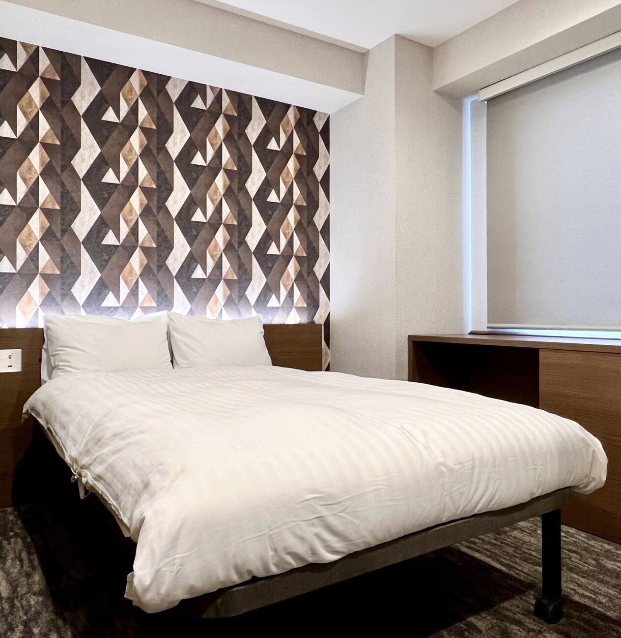 A hotel room with a bed, a geometric-patterned headboard, a window with blinds, and a wooden nightstand.
