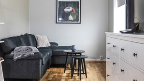 En moderne stue med en mørkegrå sofa, trægulv, et hvidt skab med skuffer, et indrammet billede af en lunsefugl og en loftslampe.
