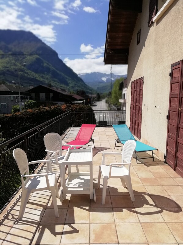 A patio with white plastic chairs and a table, a red and a blue lounge chair, and a view of mountains in the background.