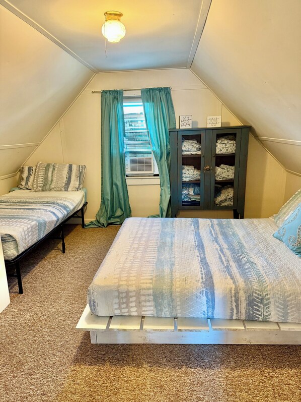 A small, triangular attic bedroom with two beds, a wardrobe, and a window with curtains. - Beach front property steps from Long Island Sound! (Westbrook)