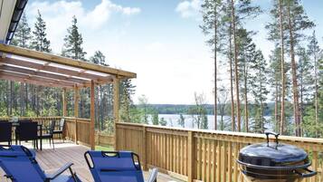 Une terrasse en bois avec deux chaises longues bleues et un barbecue, donnant sur un lac et une forêt.