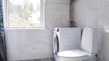 Une salle de bain équipée d’un lave-linge, de toilettes et d’une fenêtre donnant sur des arbres et un immeuble.