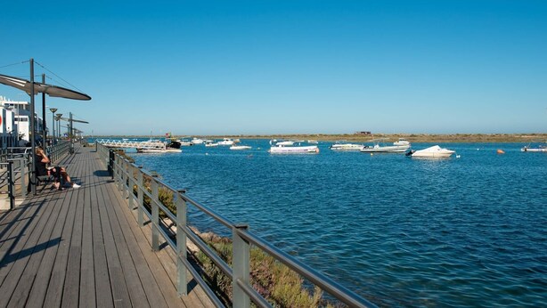 A wooden pier with a railing, boats in the water, and a clear blue sky. - Exclusive T2 apartment in the center of Cabanas de Tavira (Cabanas de Tavira)