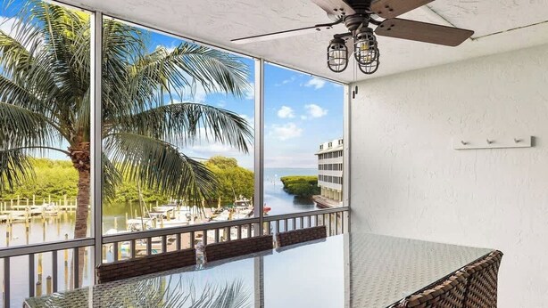 A room with a glass table, a ceiling fan, and a view of a palm tree and water. - Bayside Villa 4208 | Stunning Harbourview Residence| Walk to the Beach (Captiva)