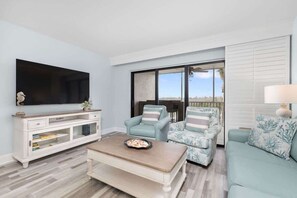 A living room with a flat-screen TV, a white entertainment unit, a coffee table, two patterned armchairs, a sofa, and a large window with a view. - Stunning Harborview Residence on Captiva Island (Captiva)