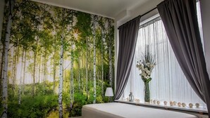 A bedroom with a large forest mural, a bed, a window with curtains, and a vase with flowers. - Lava Apartment (Vestmannaeyjar)