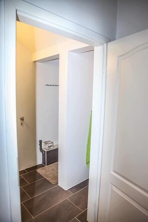 A narrow hallway with a door leading to a bathroom. The bathroom has a toilet, a sink, and a towel hanging on the wall. The floor is tiled, and the walls are painted white.