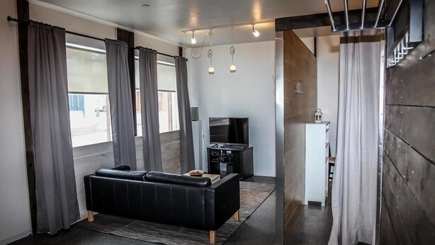 A modern living room with a black sofa, a flat-screen TV, and a small kitchen area. - Lava Apartment (Vestmannaeyjar)