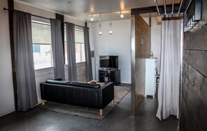 A modern living room with a black sofa, a flat-screen TV, and a small kitchen area. - Lava Apartment (Vestmannaeyjar)