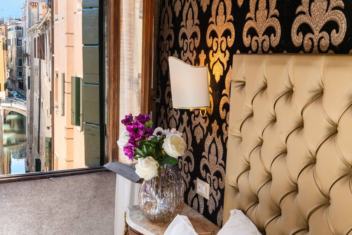 A hotel room with a bed, pillows, a vase with flowers, a lamp, and a view of a cityscape through a window.