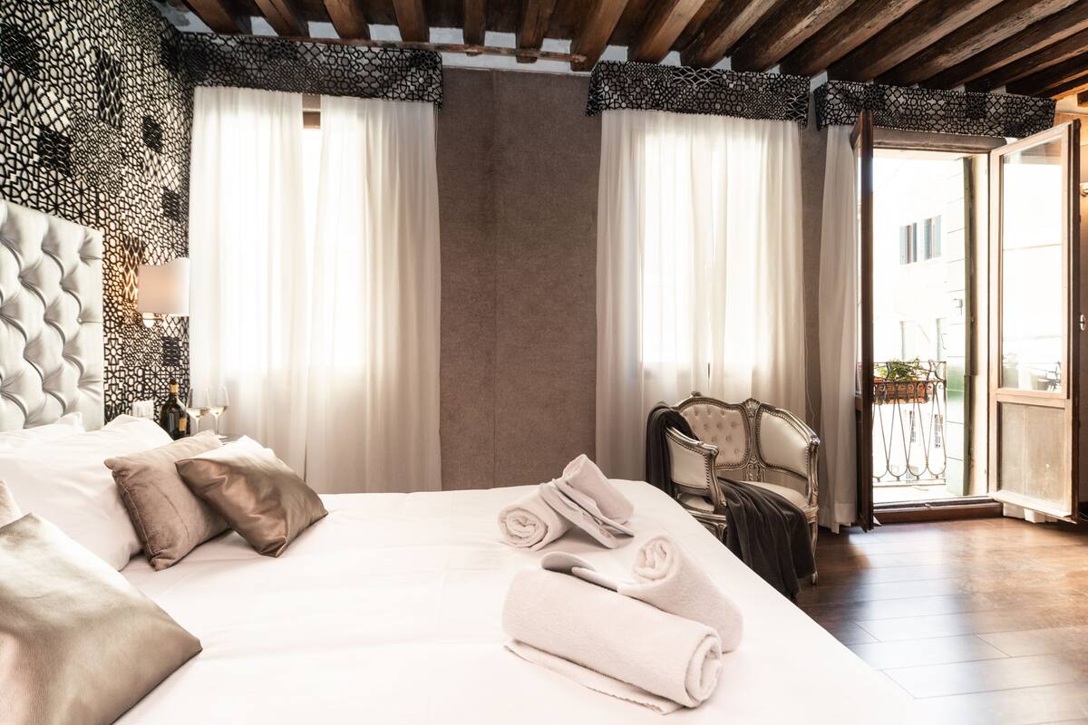 A hotel room with a large bed, a chair, a balcony with a railing, and a view of the outside.