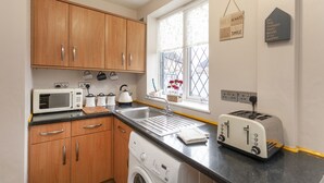 Een keuken met houten kastjes, een magnetron, een broodrooster, een wasmachine en een raam met decoratieve objecten.