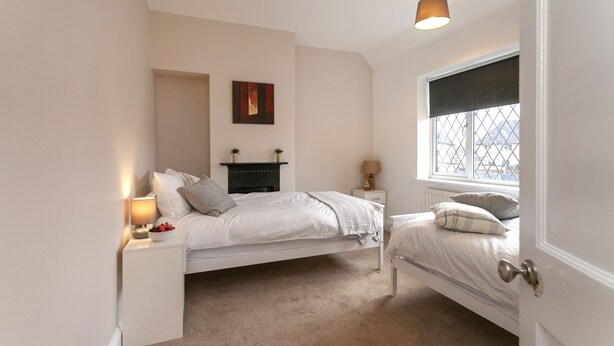 Een slaapkamer met een wit bed, een klein nachtkastje met een lamp, een raam met uitzicht en een open haard.