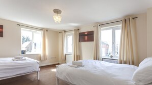 Ein Hotelzimmer mit zwei Betten, einer Chandelierleuchtre, großen Fenstern mit Vorhängen und Blick auf Gebäude.