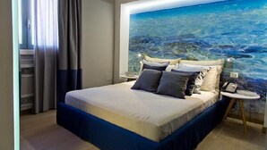 A bedroom with a bed, a nightstand, a painting of the ocean, and a window with curtains. - Masseria Sant'Eleuterio (Collepasso)