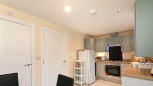 A compact kitchen with white cabinets, a built-in microwave, a stove, and a refrigerator. There is a small shelf unit and a dining table with chairs. - Condron 3 bedroom house (Worksop)
