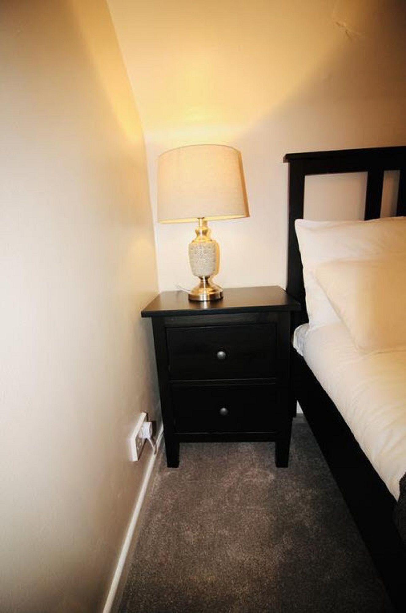 Een klein, donker houten nachtkastje met een lamp en één lade, geplaatst naast een bed met witte beddengoed.