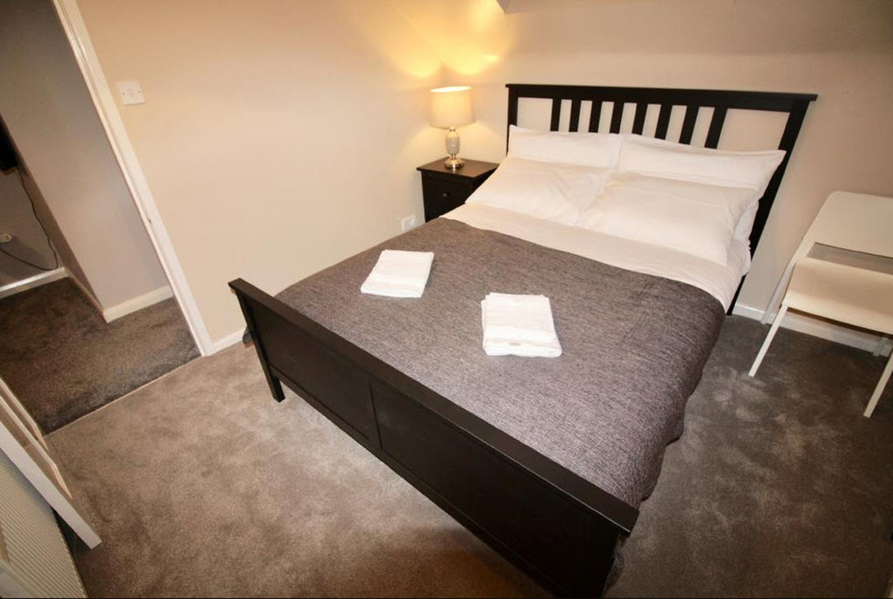 A neatly made bed with a dark headboard, a bedside lamp, and a white chair.