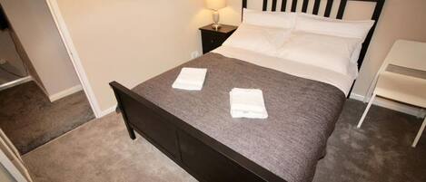 A neatly made bed with a dark headboard, a bedside lamp, and a white chair.