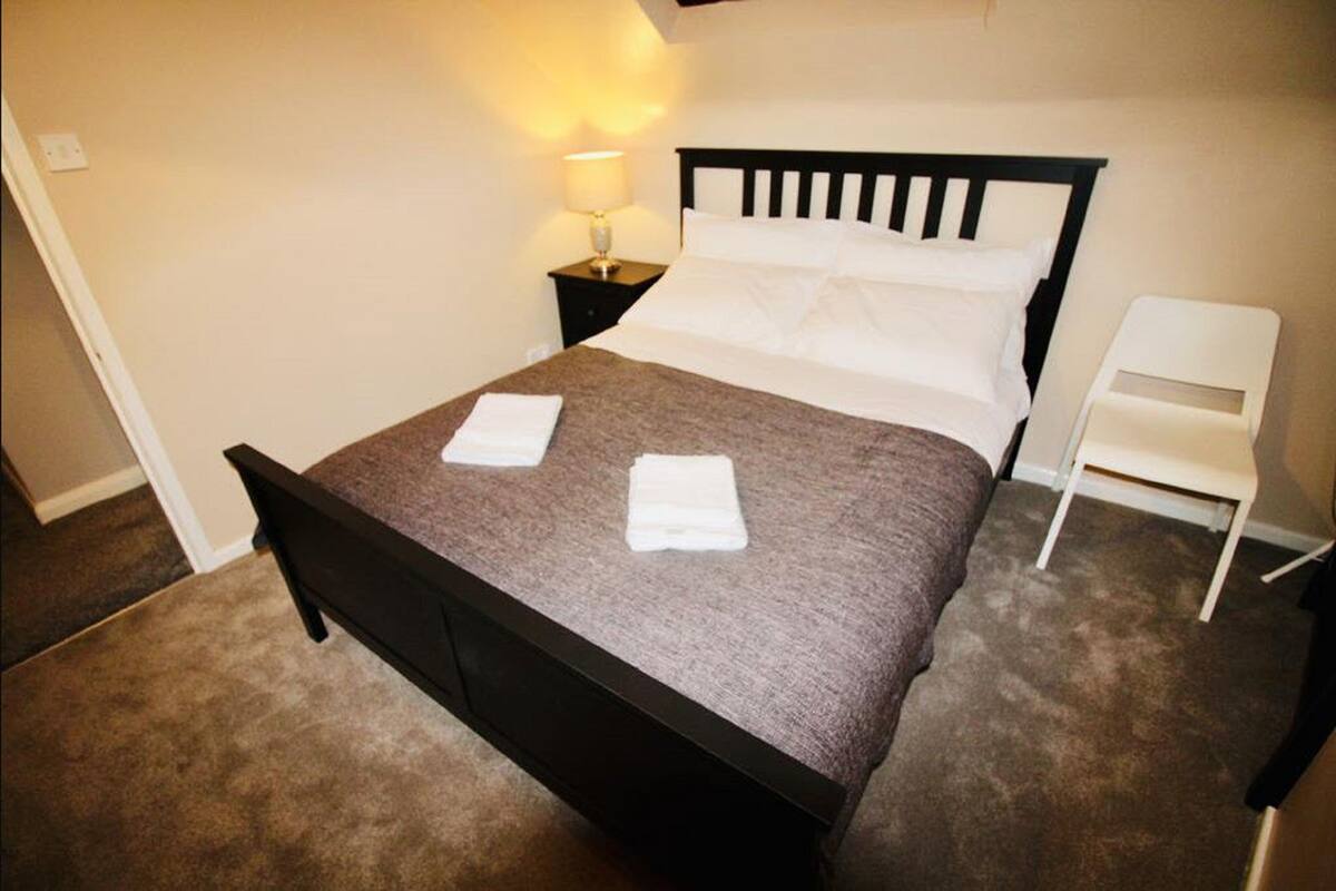 A neatly made bed with a dark headboard, a bedside lamp, and a white chair.