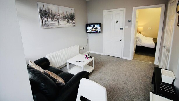 A compact living room with a black leather sofa, a white coffee table, and a television mounted on the wall. A door leads to a bedroom with a bed.