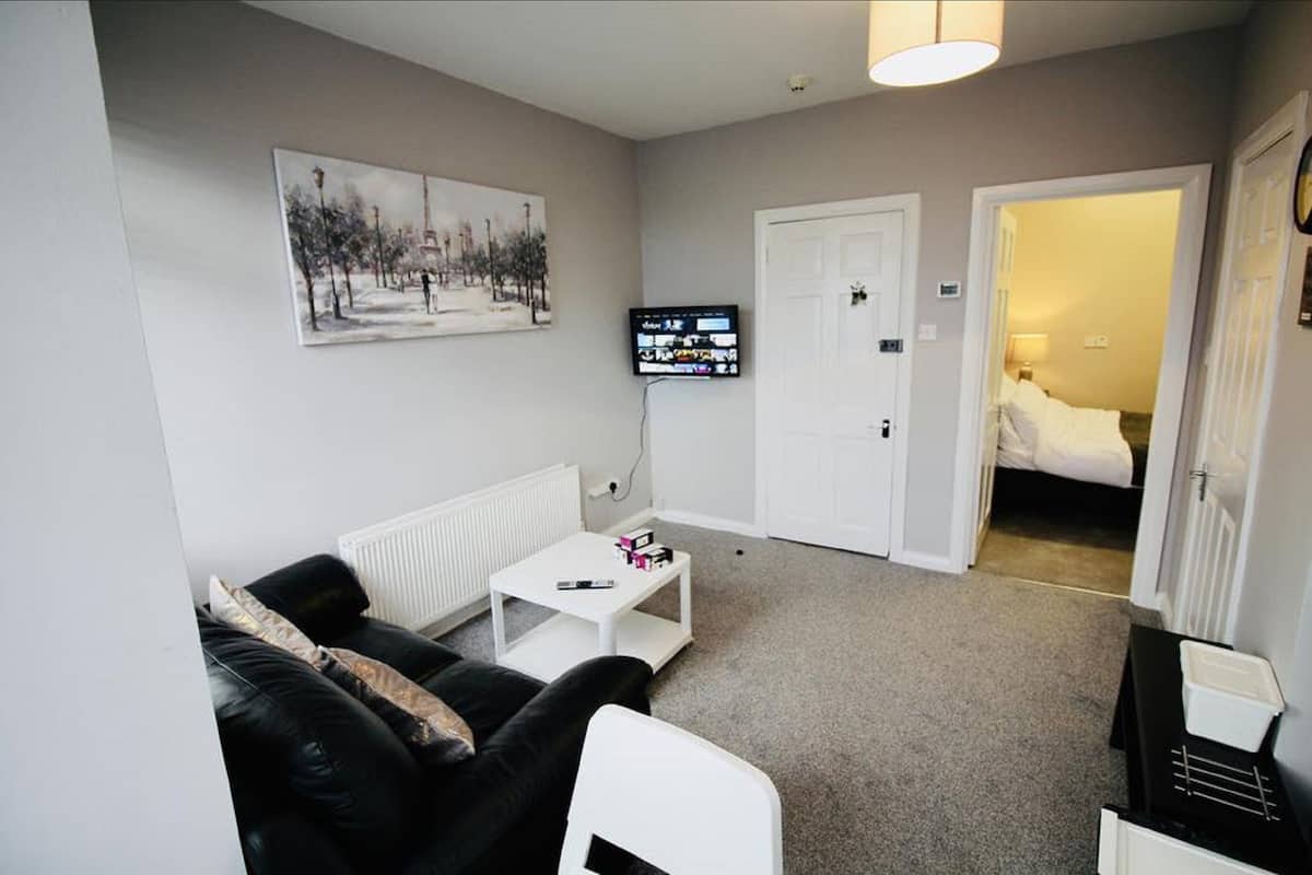 A compact living room with a black leather sofa, a white coffee table, and a television mounted on the wall. A door leads to a bedroom with a bed.