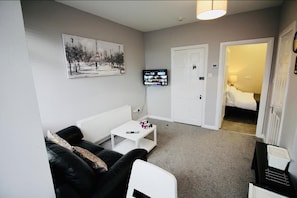 A compact living room with a black leather sofa, a white coffee table, and a television mounted on the wall. A door leads to a bedroom with a bed. - Woodstock A 1 bed apartment (Belfast)