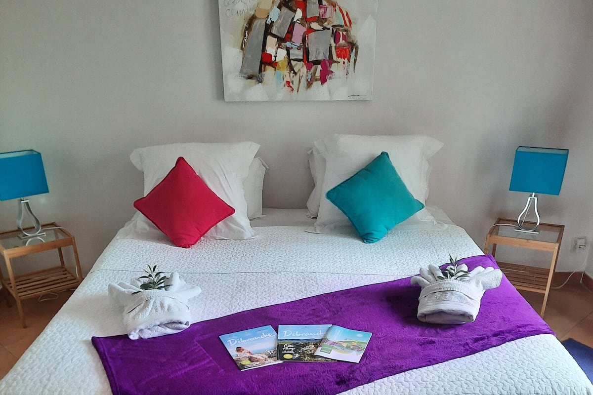 A hotel room with a bed, two nightstands, a purple throw blanket, two purple ottomans, and a painting on the wall.