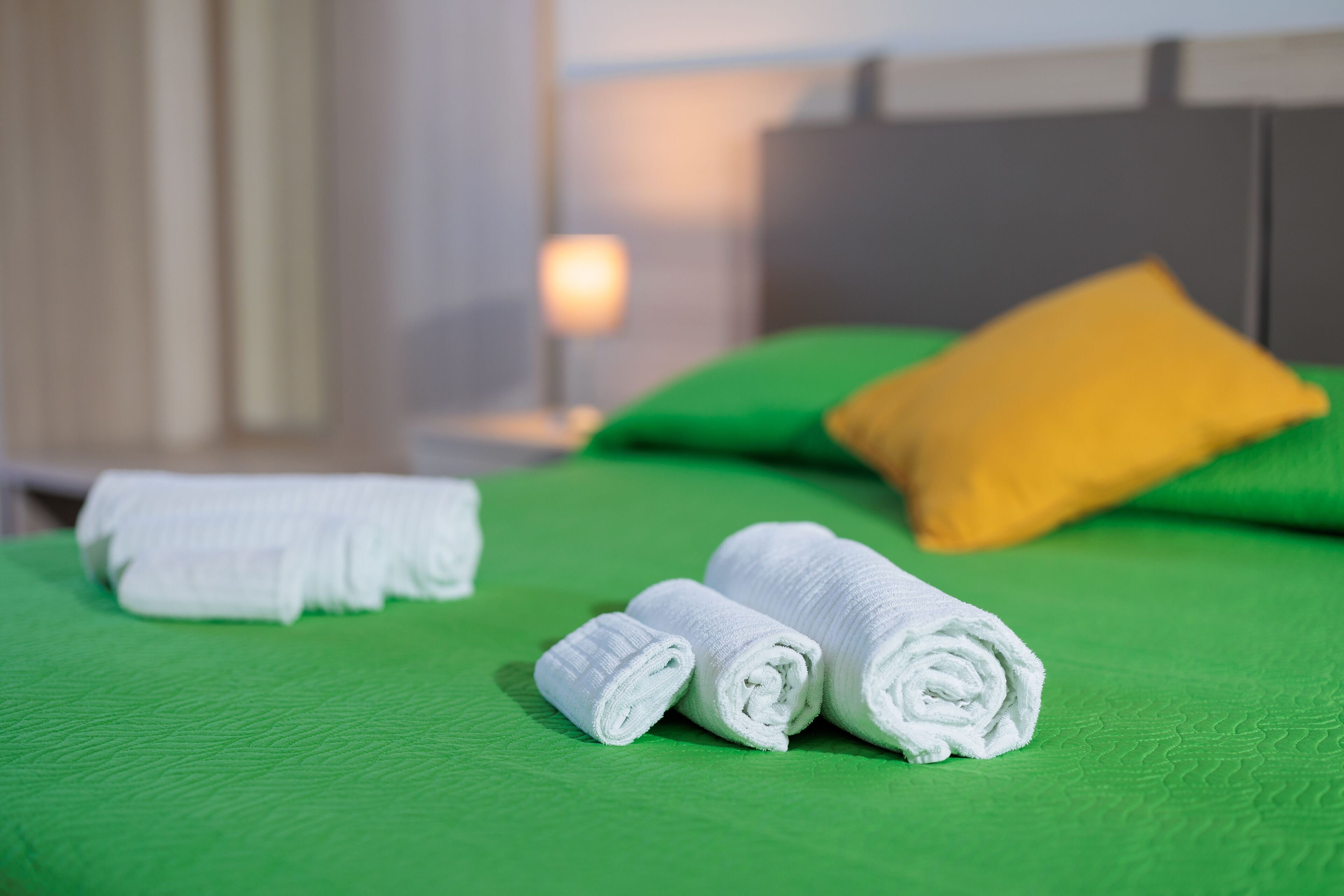A neatly made bed with green and yellow pillows, and rolled white towels.
