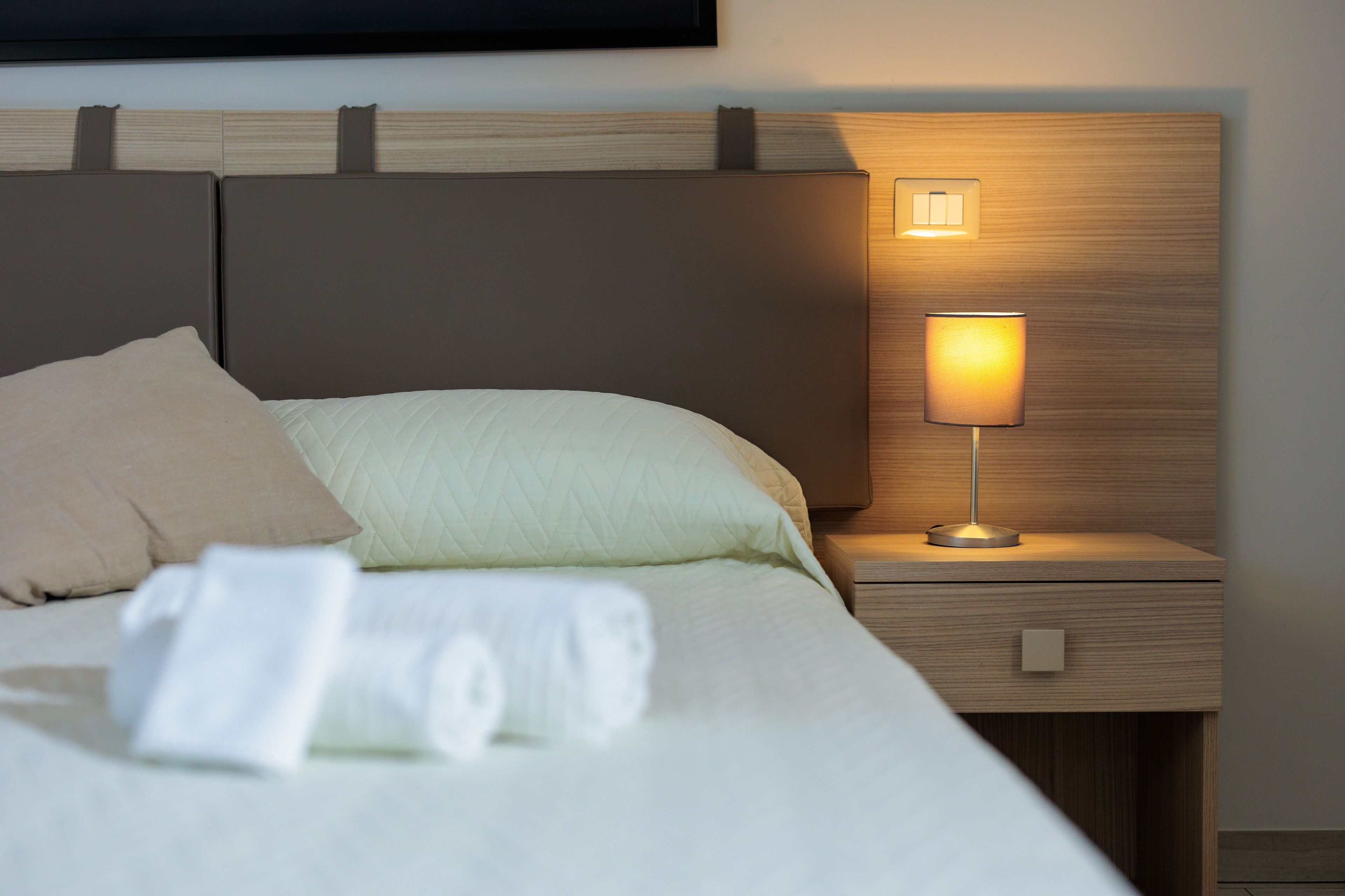 Cama bem arrumada com travesseiro e toalha enrolada, abajur e mesa de cabeceira de madeira.