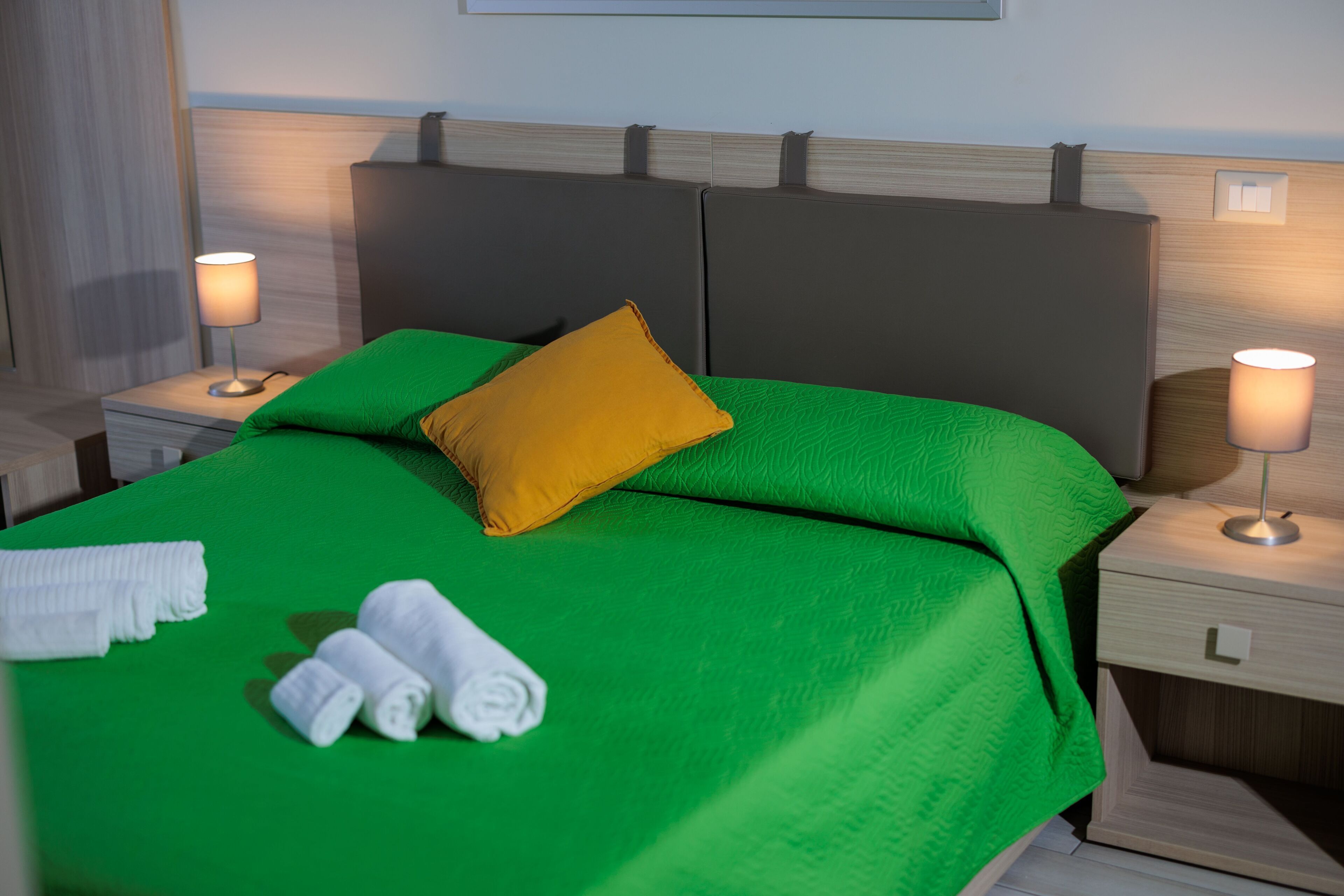 A neatly made bed with green bedding and a yellow pillow, two bedside lamps, and neatly folded white towels on the nightstand.