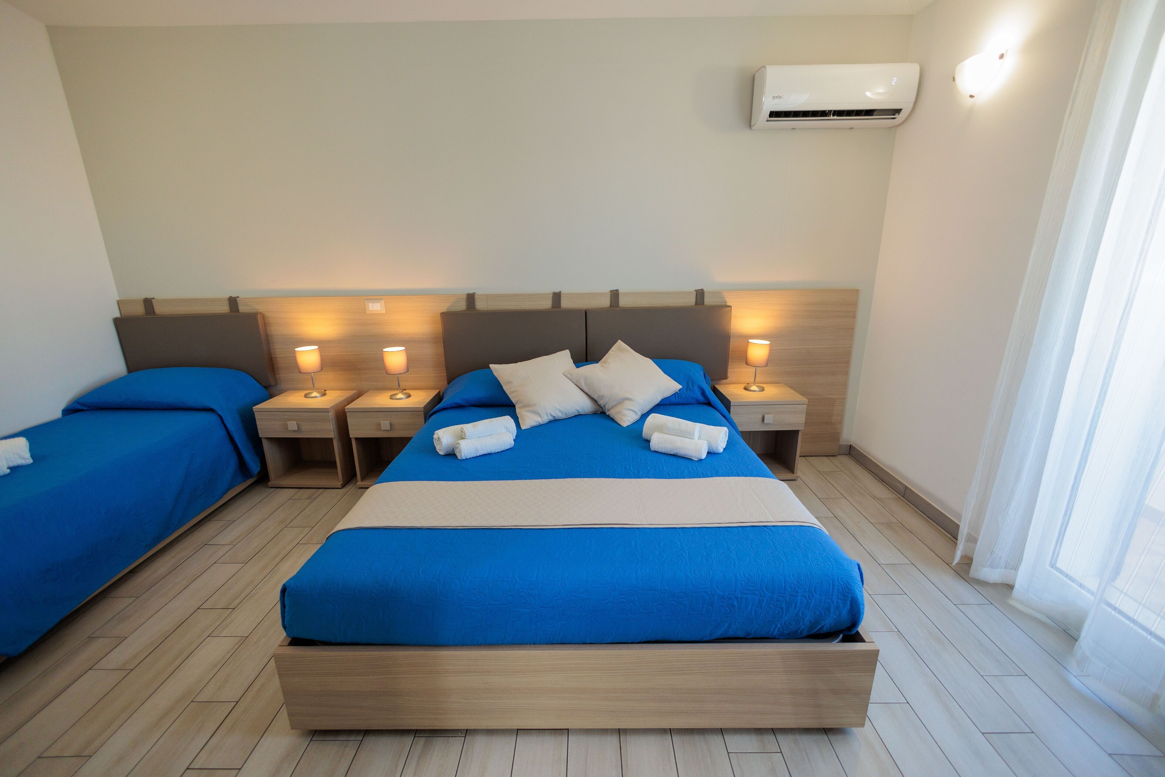A hotel room with a large bed, two bedside tables with lamps, a headboard, and an air conditioning unit.