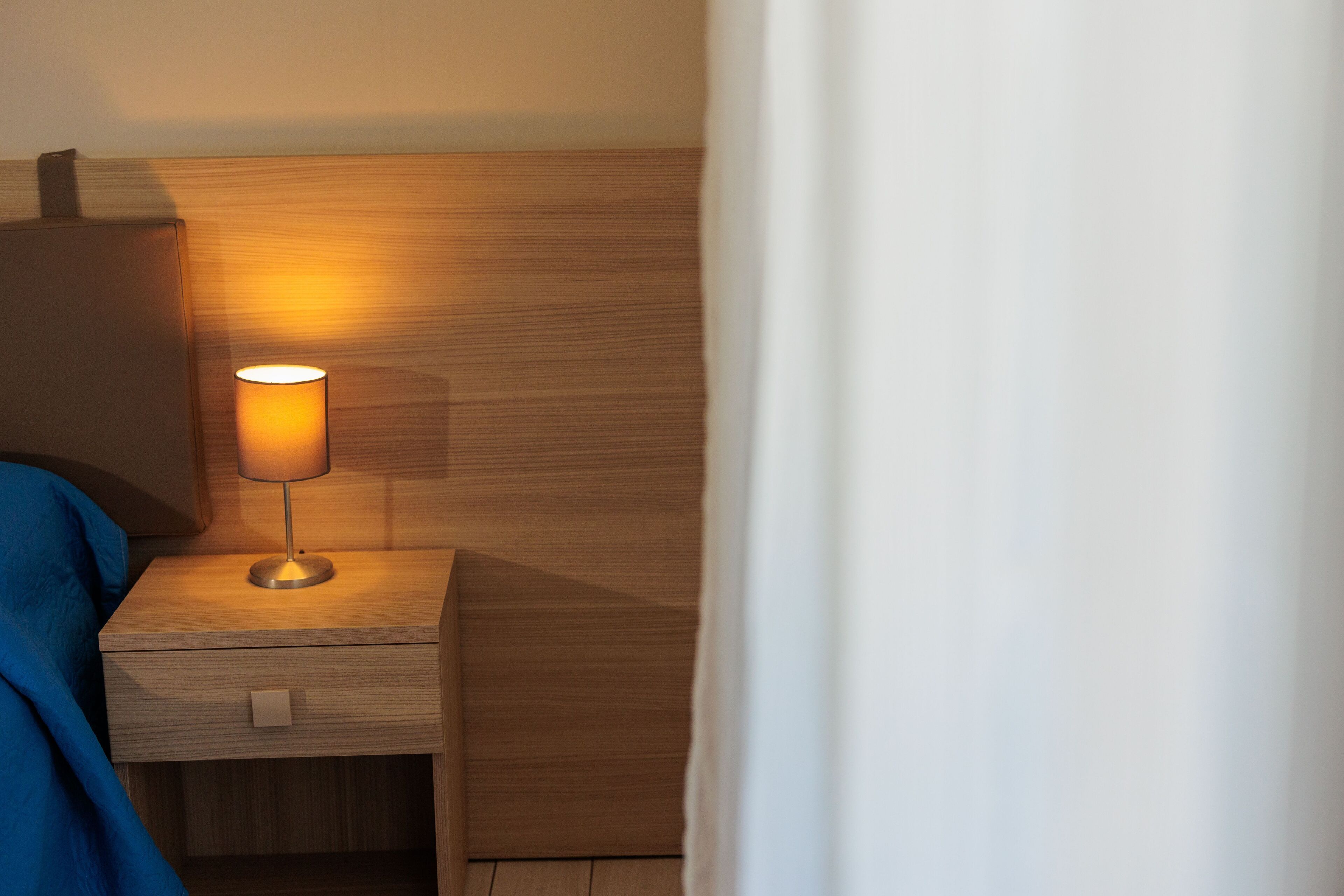 A wooden bedside table with a lamp, a drawer, and a blue blanket.
