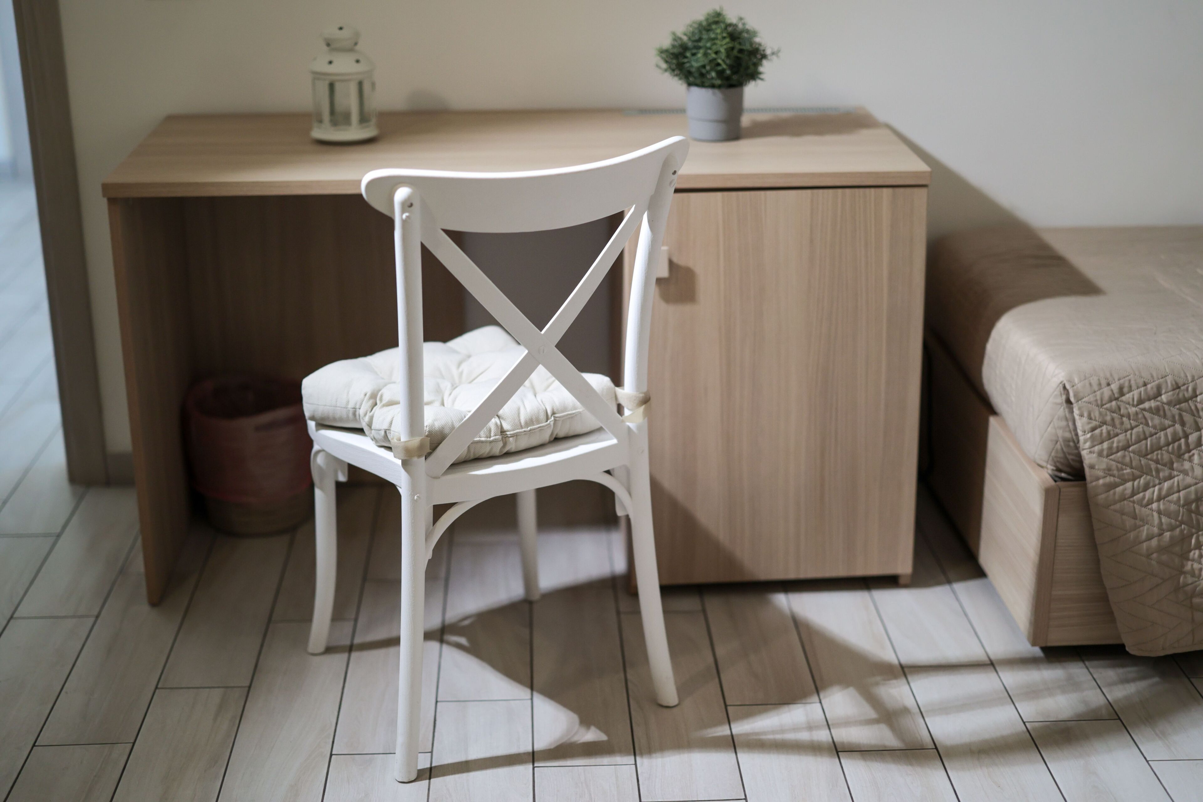 Ein ordentlich eingerichtetes Schlafzimmer mit einem Holztisch, einem weißen Stuhl, einem Bett mit einer beige farbenen Steudecke und einem kleinen Topfpflanzen auf dem Tisch.