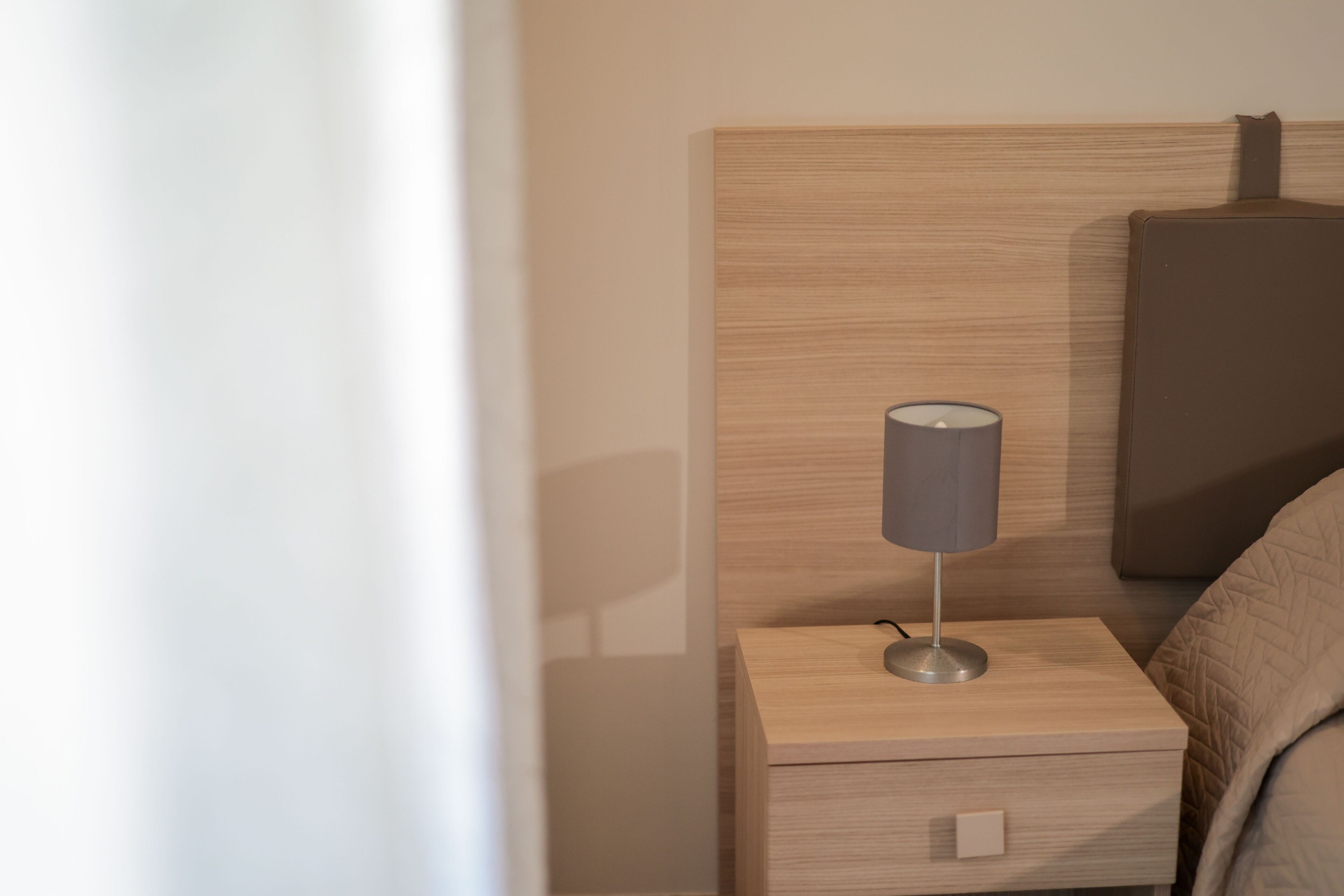 Uma mesinha de cabeceira de madeira bem arrumada, com uma luminária, uma gaveta e uma cama com cobertura acolchoada.