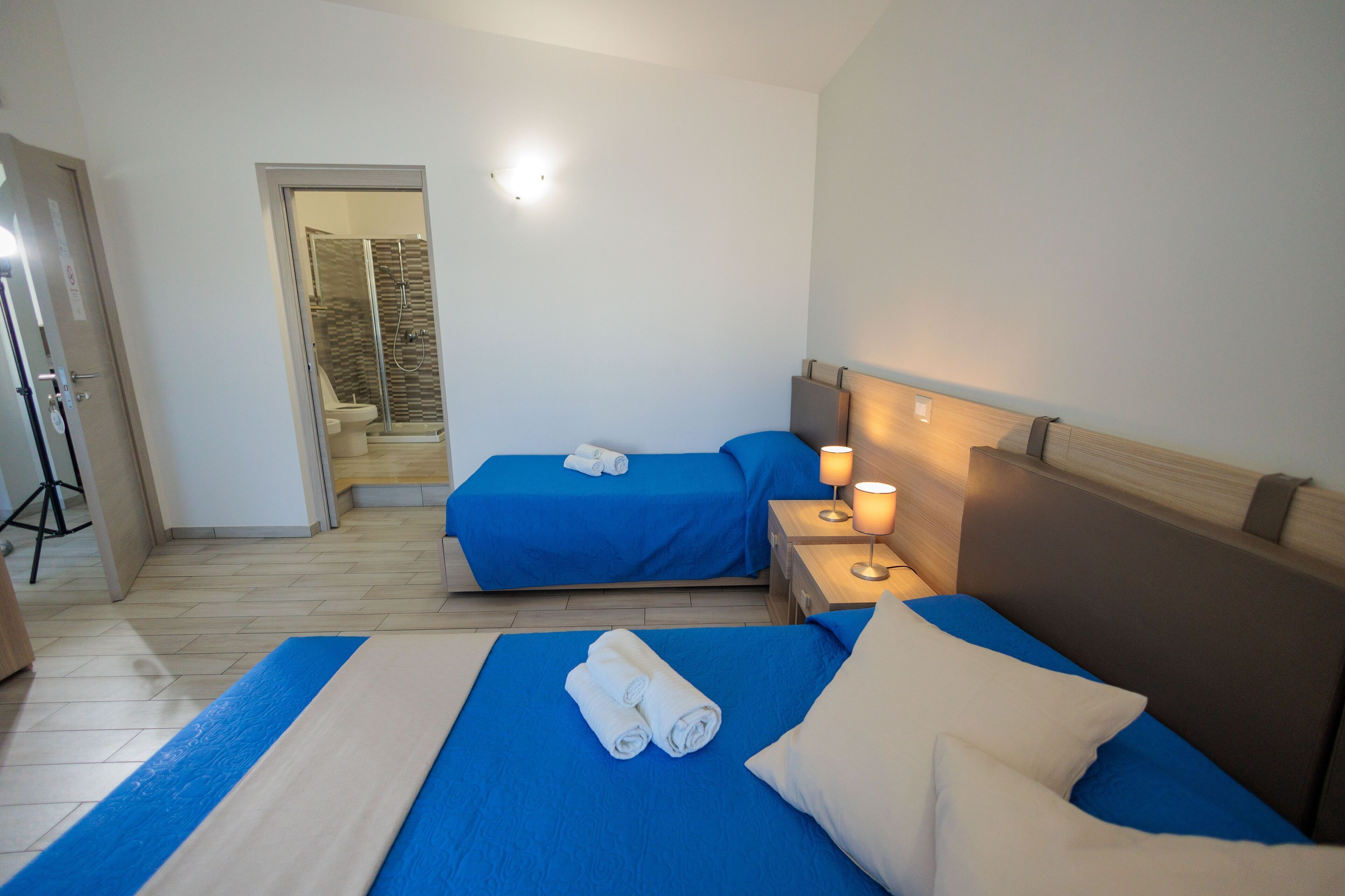 A hotel room with two beds, each with blue bedding and a white pillow, a bedside table with a lamp, and a shower area visible through a glass door.