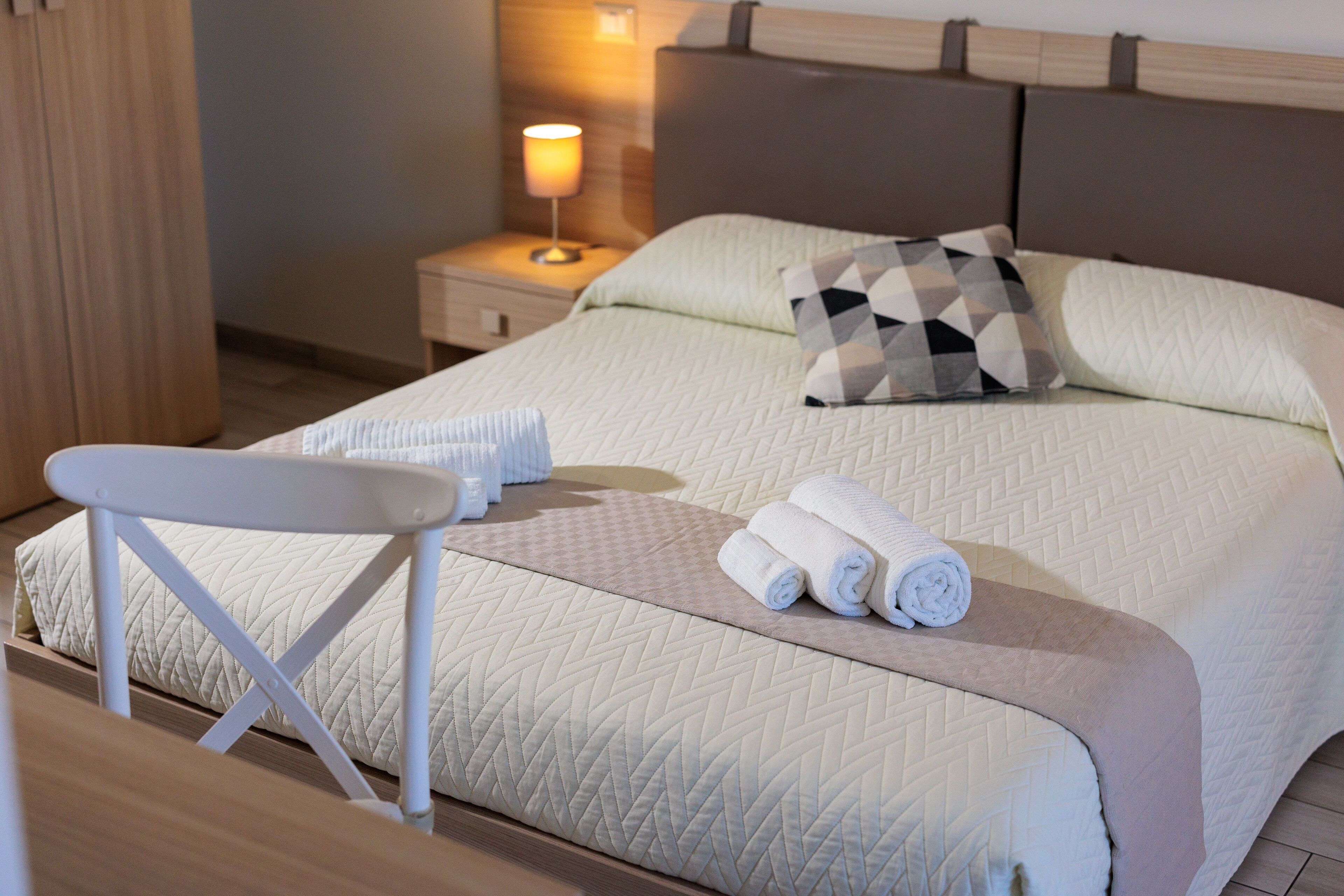 A neatly made bed with a quilted cover, a pillow, and rolled towels on the bed. There is a small table with a white chair beside the bed.