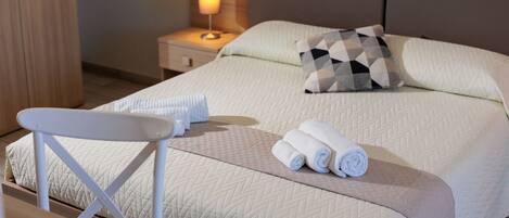 A neatly made bed with a quilted cover, a pillow, and rolled towels on the bed. There is a small table with a white chair beside the bed.